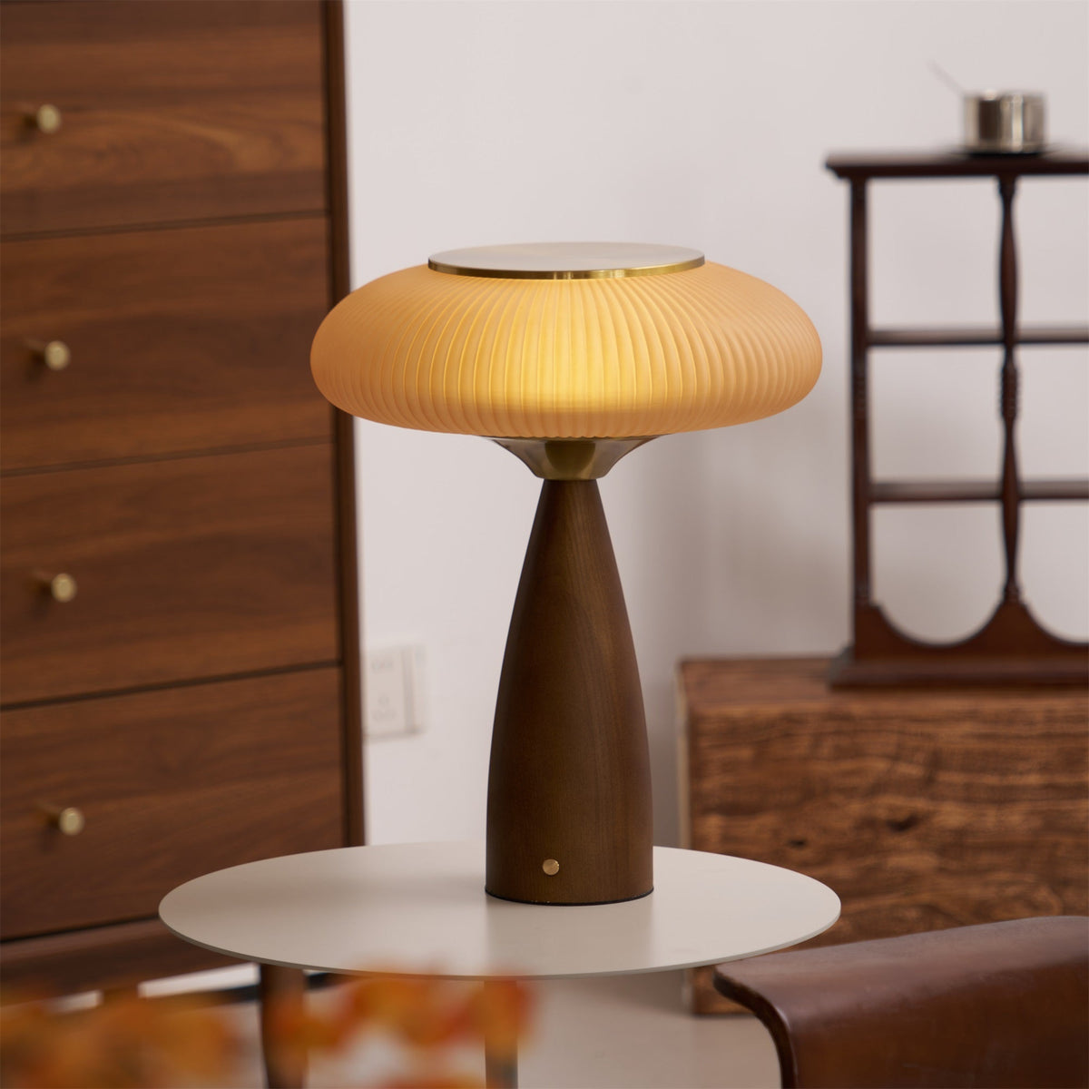 Lit cinnamon shade of this Clérachic table lamp on a small white round table, with a tall walnut chest of drawers behind.