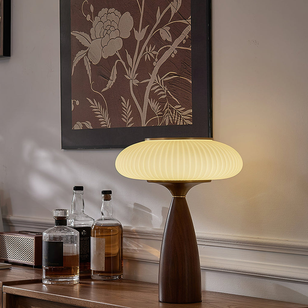 Lit ribbed shade of this Clérachic lamp on a wooden sideboard under a botanical print, with whisky bottles at its left.