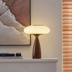 A Clérachic table lamp glowing on a white ledge beside stacked books, with a landscape painting and wooden blinds behind.