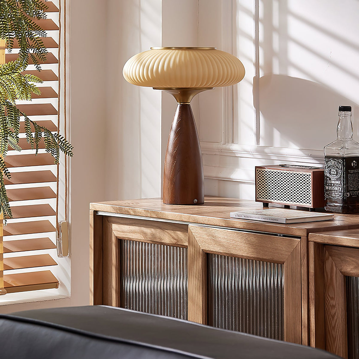 This Clérachic lamp on a slatted-glass wooden sideboard beside a retro speaker and a bottle, with sunlit blinds at the window.
