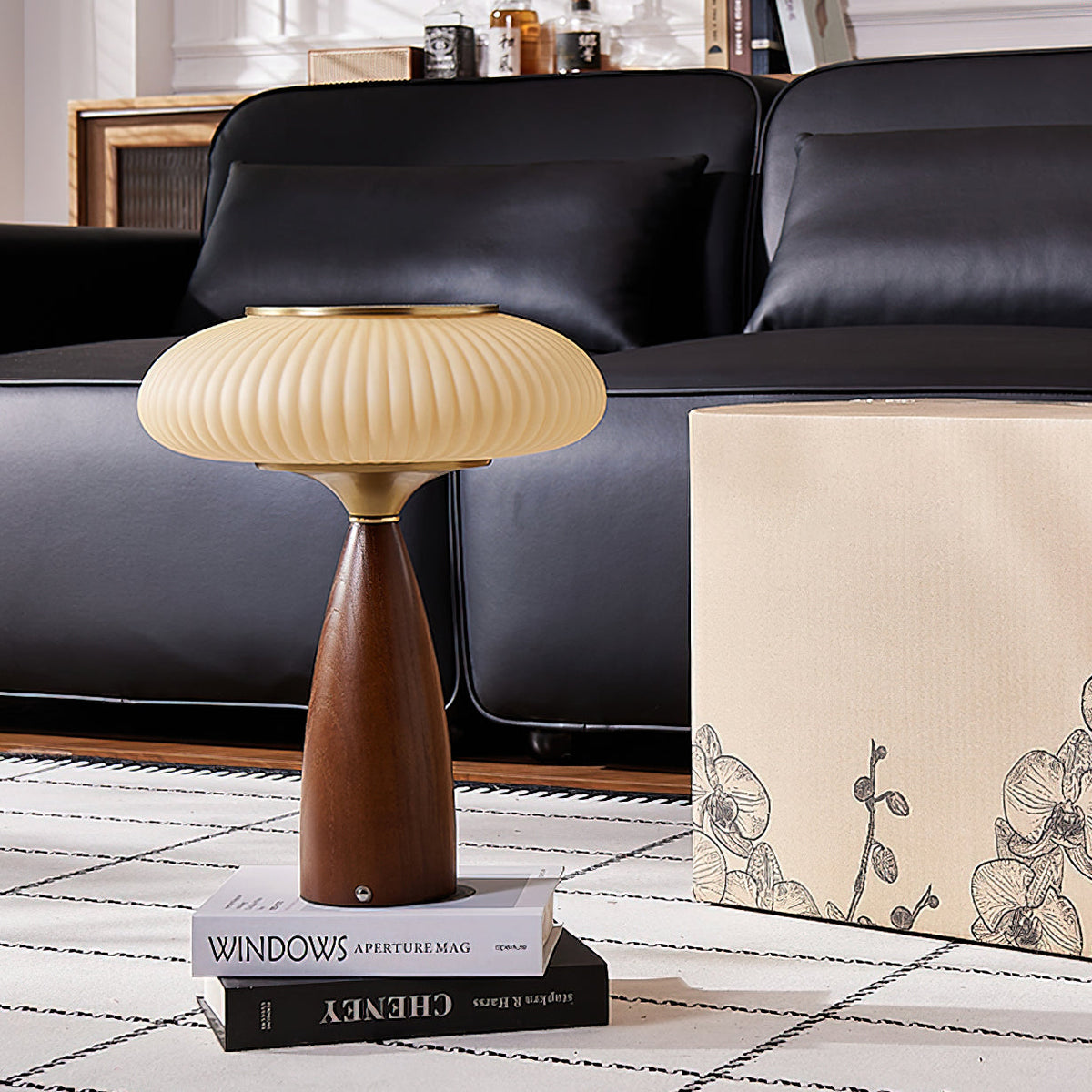 This Clérachic lamp stands on a pair of books on a patterned rug, in front of a black leather sofa and a floral pouffe.