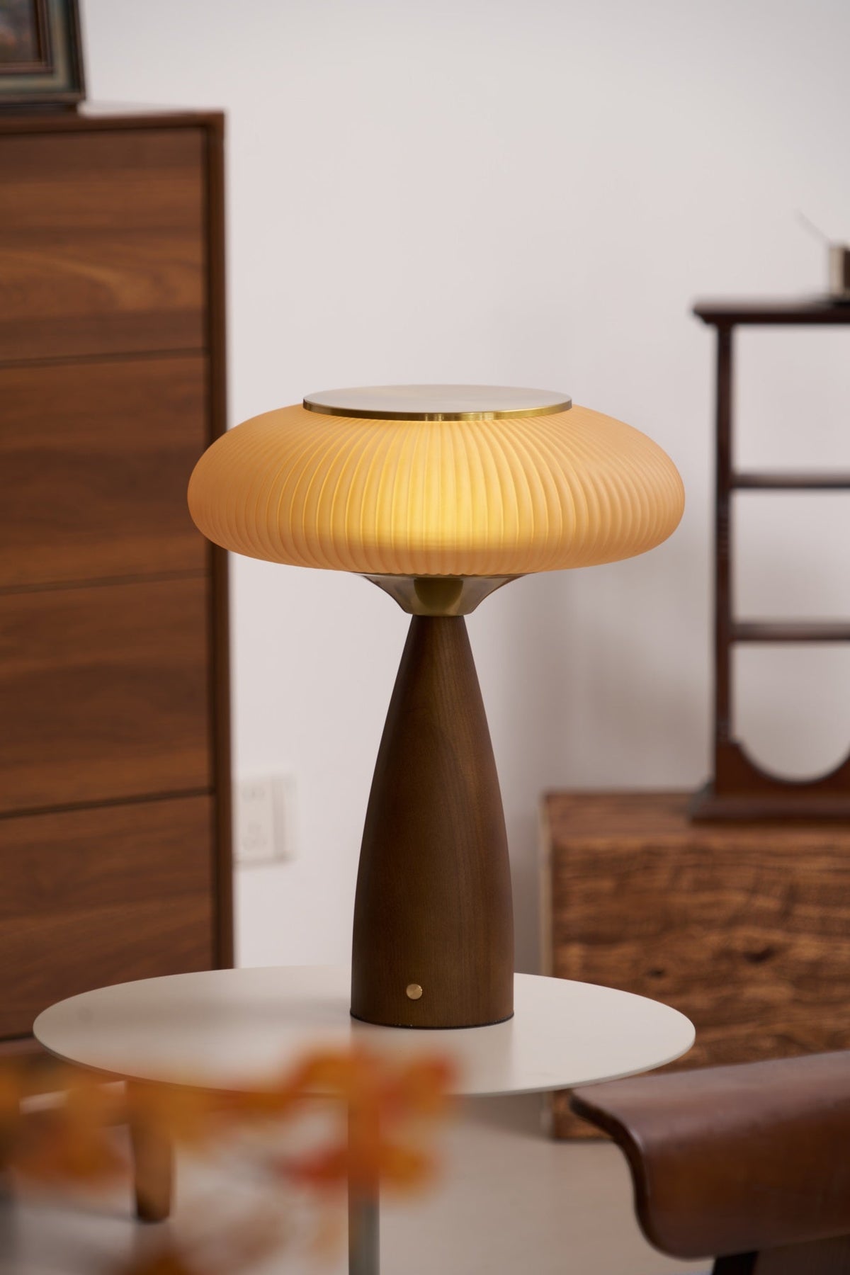 Lit Clérachic table lamp on a small white round table beside a tall chest of drawers, with orange leaves at the front.
