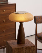 Lit Clérachic ribbed lamp on a rough wooden block, with a walnut chest of drawers and a woven-fabric chair beside it.