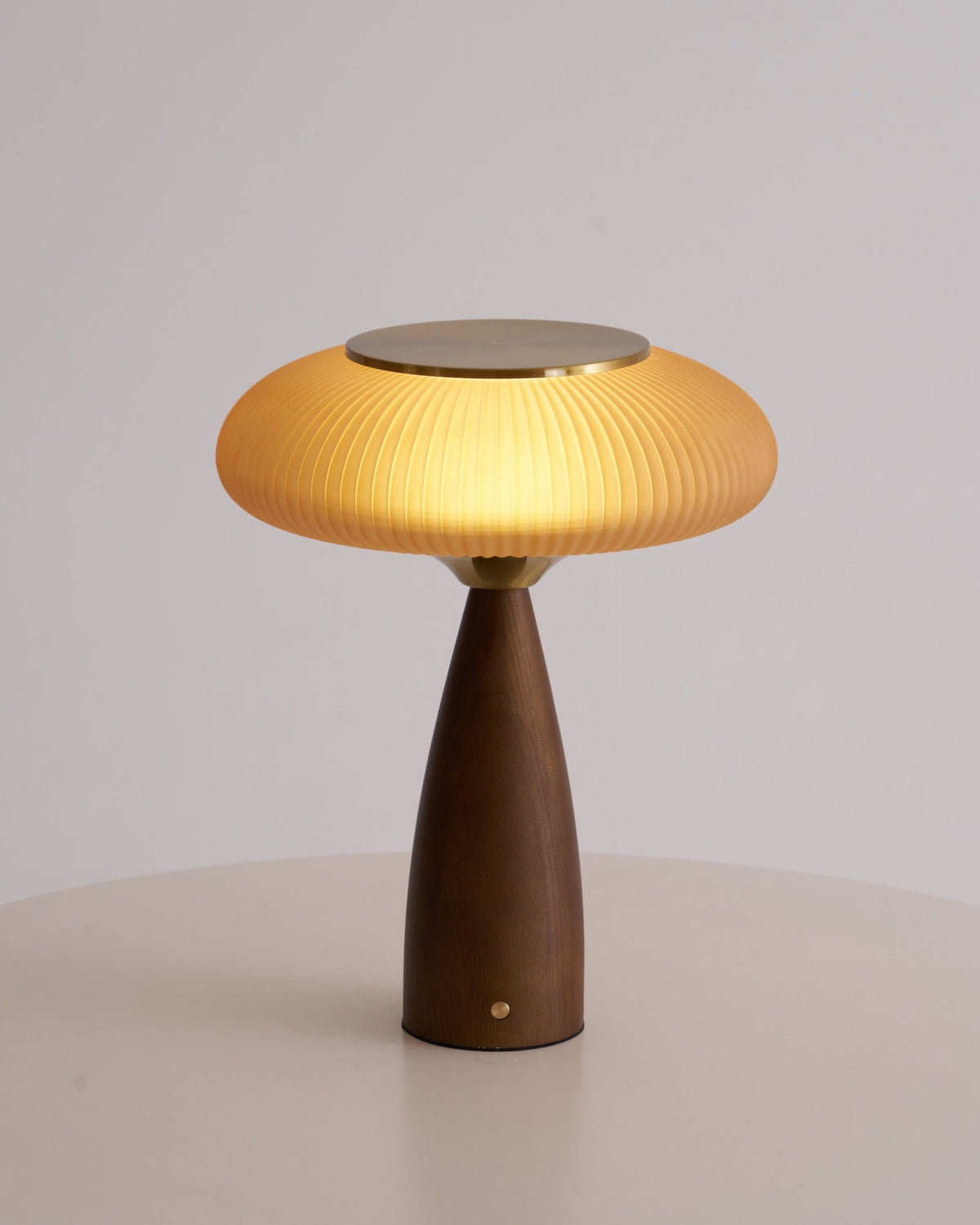 This Clérachic table lamp, lit, on a round white table against a pale grey wall, its cinnamon shade glowing warmly.