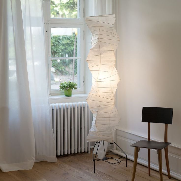 Clérachic tall faceted paper-lantern floor lamp lit beside white curtains and a radiator, with a dark wooden chair standing to its right.