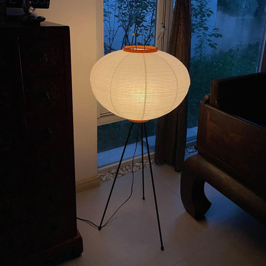 Clérachic oval rice-paper floor lamp lit after dark between a carved antique cabinet and a wooden daybed, the window behind it deep blue.