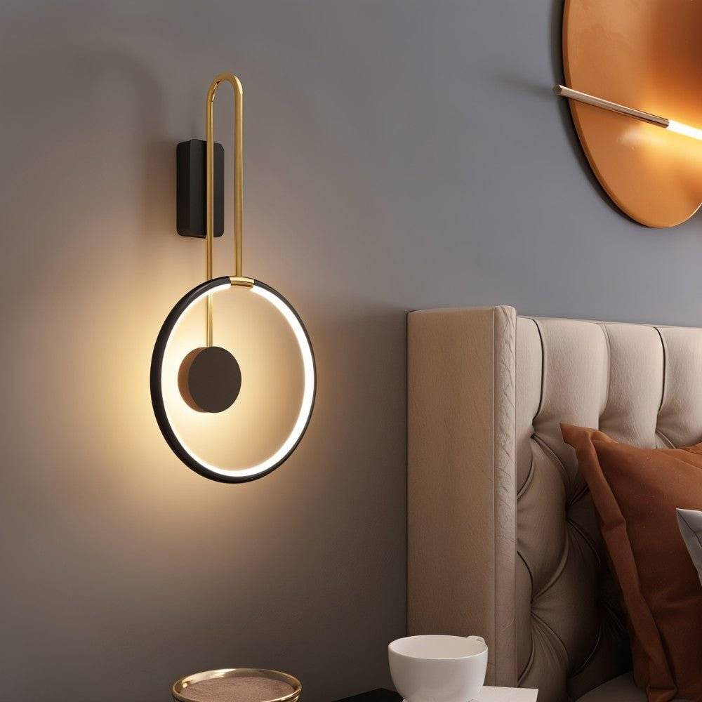 Clérachic black ring wall light with a brass-toned arm glowing warm beside a tufted headboard in a grey bedroom.