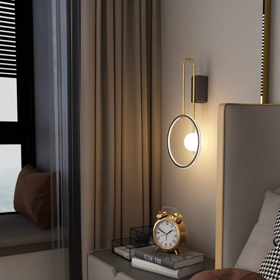 Clérachic ring wall light with a glass ball and brass-toned arm, glowing beside curtains and a grey upholstered headboard.
