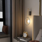 Clérachic ring wall light with a glass ball and brass-toned arm, glowing beside curtains and a grey upholstered headboard.