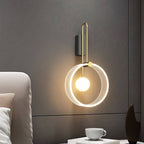 Clérachic wall light with a clear fluted acrylic ring and glowing glass ball on a brass-toned arm, against a textured grey wall.