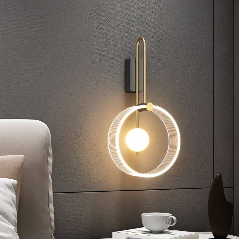 Clérachic wall light with a clear fluted acrylic ring and glowing glass ball on a brass-toned arm, against a textured grey wall.