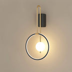 Clérachic black ring wall light with a brass-toned arm and a lit white glass ball, mounted on a plain warm taupe wall.
