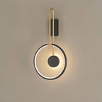 Clérachic black ring wall light with a brass-toned U-shaped arm and a black disc, glowing warm on a taupe wall.