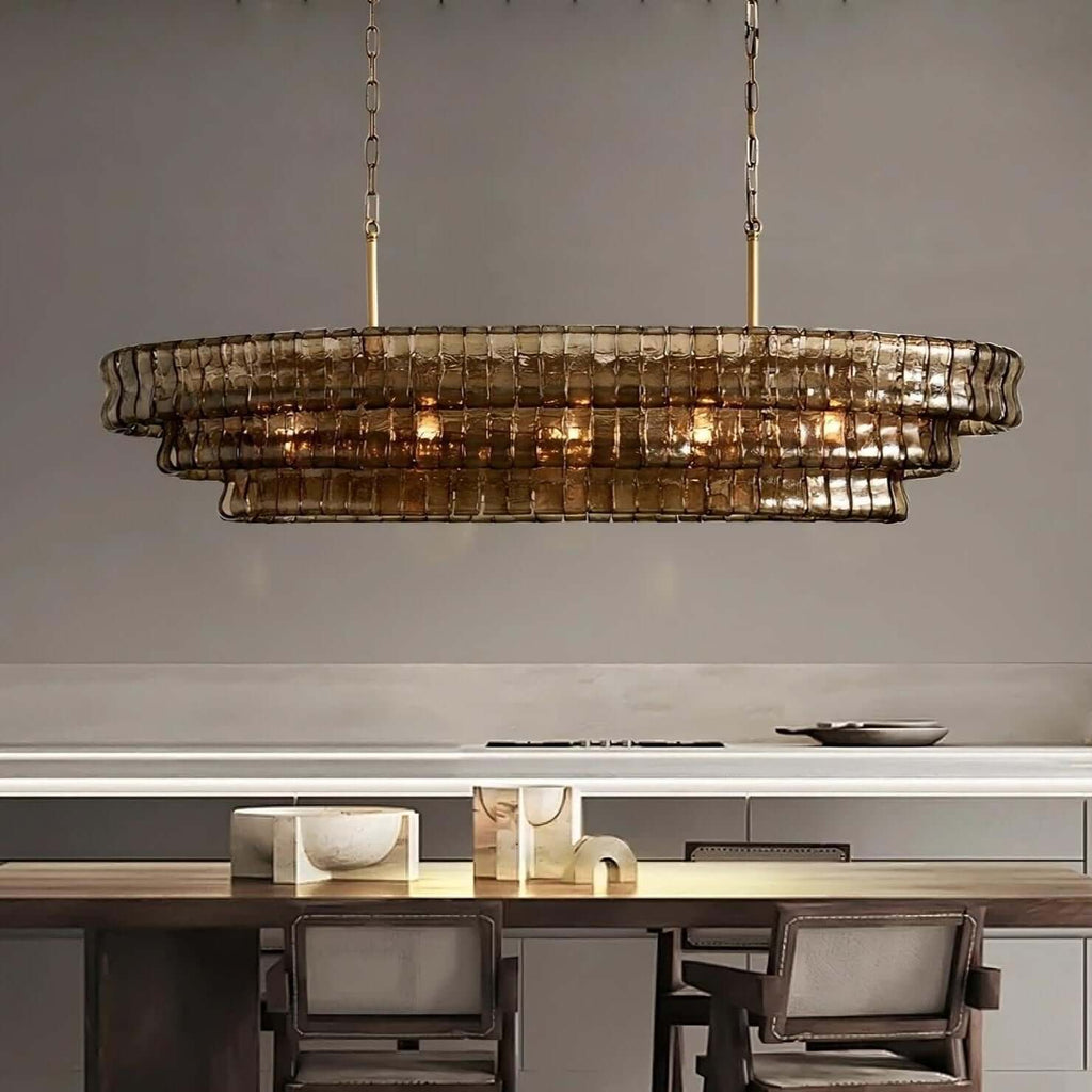 Smoke-brown stepped glass tiers of this Clérachic chandelier, lit warm, hanging above a dark wood table by a pale kitchen counter.