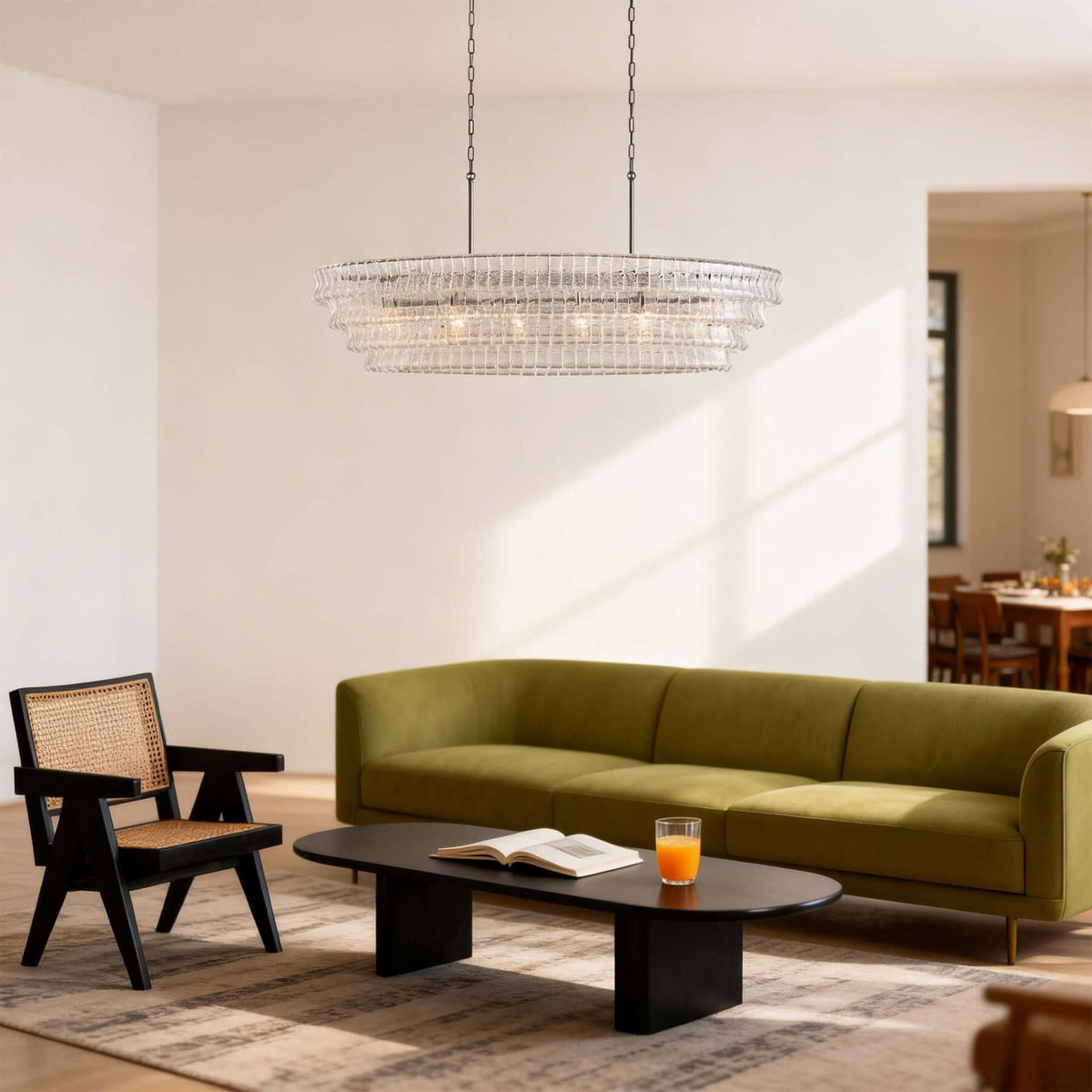 A Clérachic linear chandelier in clear glass tiers hangs in a bright living room with an olive-green sofa and a low black coffee table.