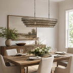Above a rustic dining table set for dinner, this Clérachic chandelier shows layered grey-tinted glass beside a landscape painting.