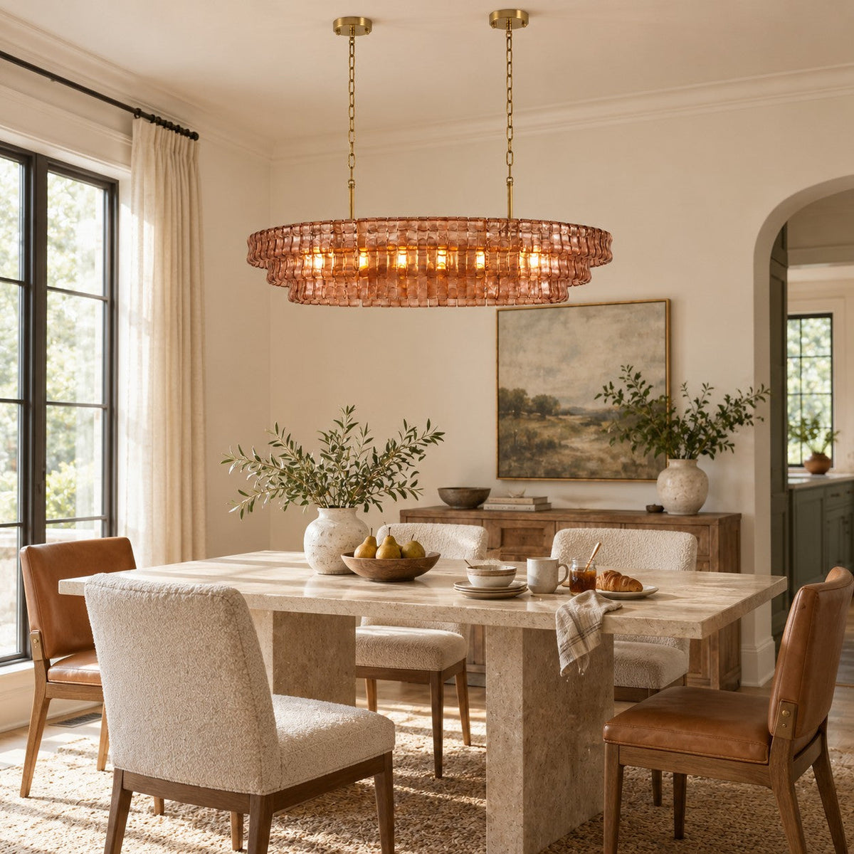 A rose-pink Clérachic chandelier with gold stems glows over a stone dining table with cream armchairs and tan leather chairs.