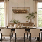 A long, dark-framed Clérachic chandelier with lit glass tiers hangs above a dark dining table and cream upholstered chairs.
