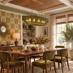 A green-glass Clérachic chandelier lights a colourful dining room with olive velvet chairs, patterned wallpaper and a wooden table.