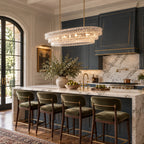 This Clérachic chandelier in clear glass hangs over a marble island with green velvet stools, in front of navy kitchen cabinets.