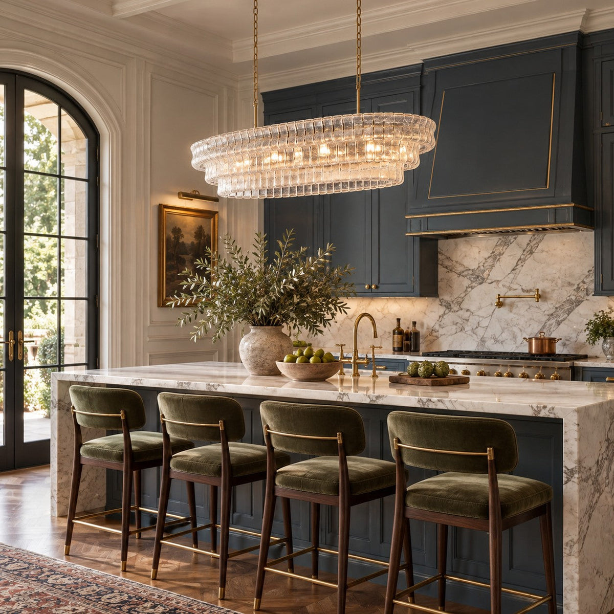 This Clérachic chandelier in clear glass hangs over a marble island with green velvet stools, in front of navy kitchen cabinets.