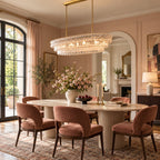 A Clérachic chandelier with gold stems and clear glass hangs over a pale oval table in a pink room with an arched doorway.