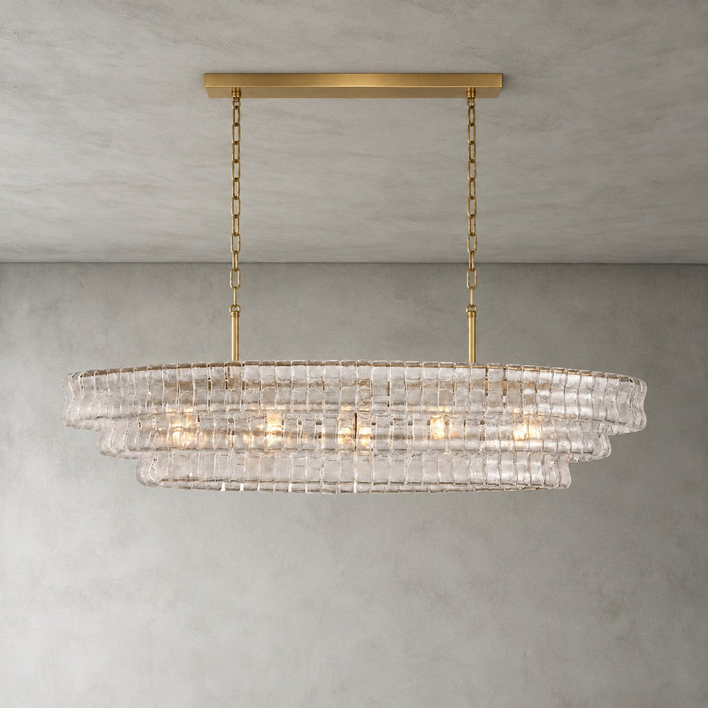 Clear glass tiers of this Clérachic chandelier, held by a gold long canopy bar and chains, lit on a grey wall.