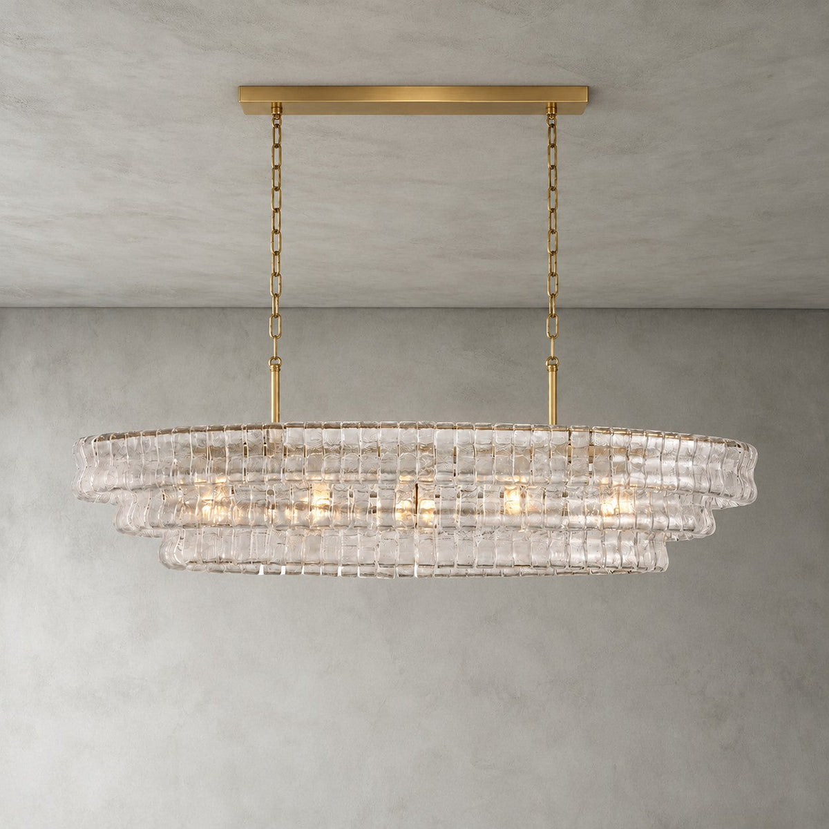 Clear glass tiers of this Clérachic chandelier, held by a gold long canopy bar and chains, lit on a grey wall.