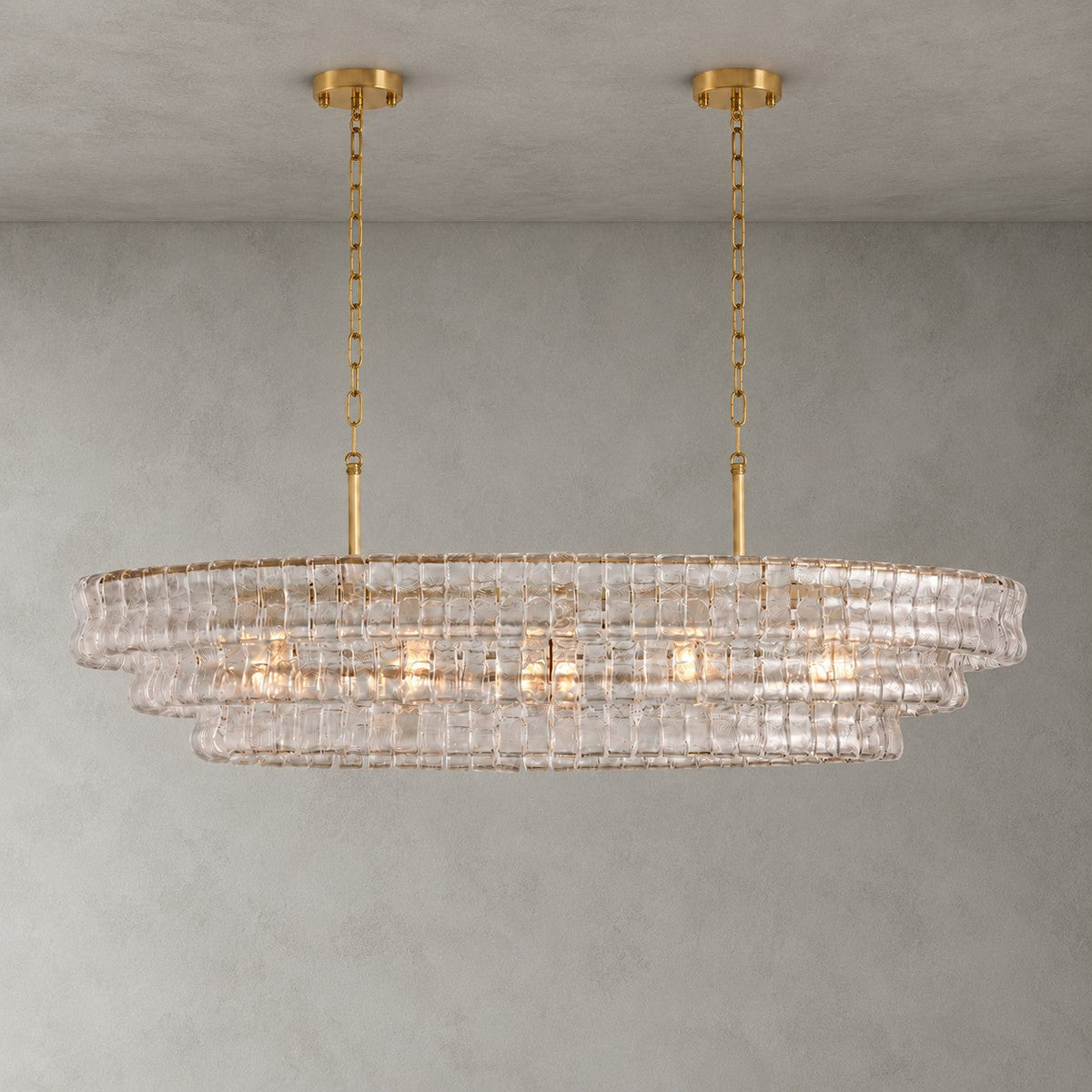 This Clérachic chandelier in clear glass hangs from two gold round canopies, its stepped tiers lit against a grey wall.