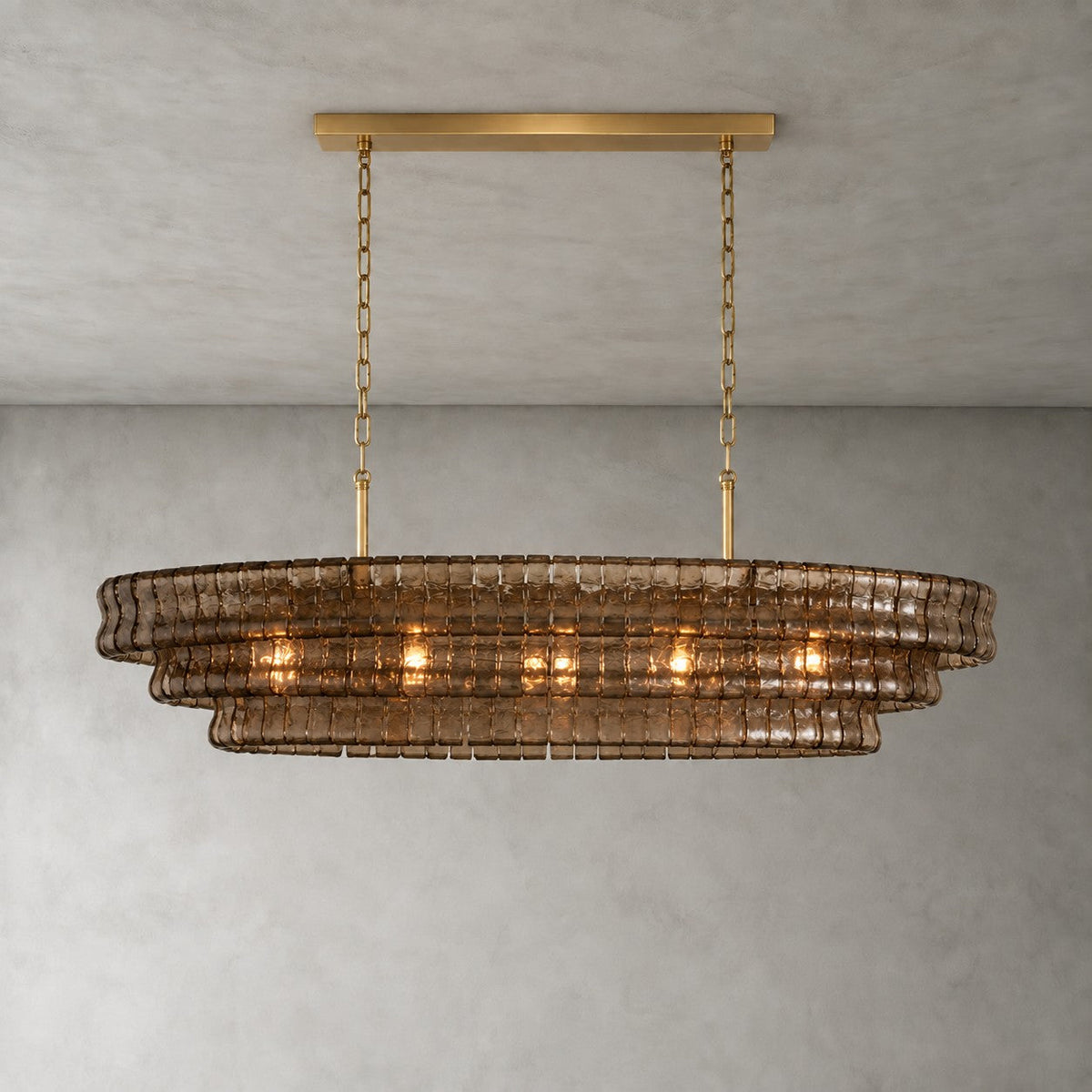 Smoky brown glass tiers on a Clérachic chandelier held by a gold long canopy bar, shown lit against a grey wall.