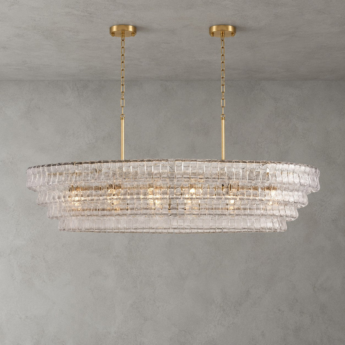 Clear glass on a four-tier Clérachic chandelier with two gold round canopies and slim stems, lit on a grey wall.