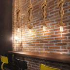 Row of Clérachic hemp rope pendants with big knots and filament bulbs lighting a red brick wall above a timber counter.