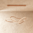 Clérachic chandelier in a warm rose gold finish, its looping rings glowing softly against a plaster wall.
