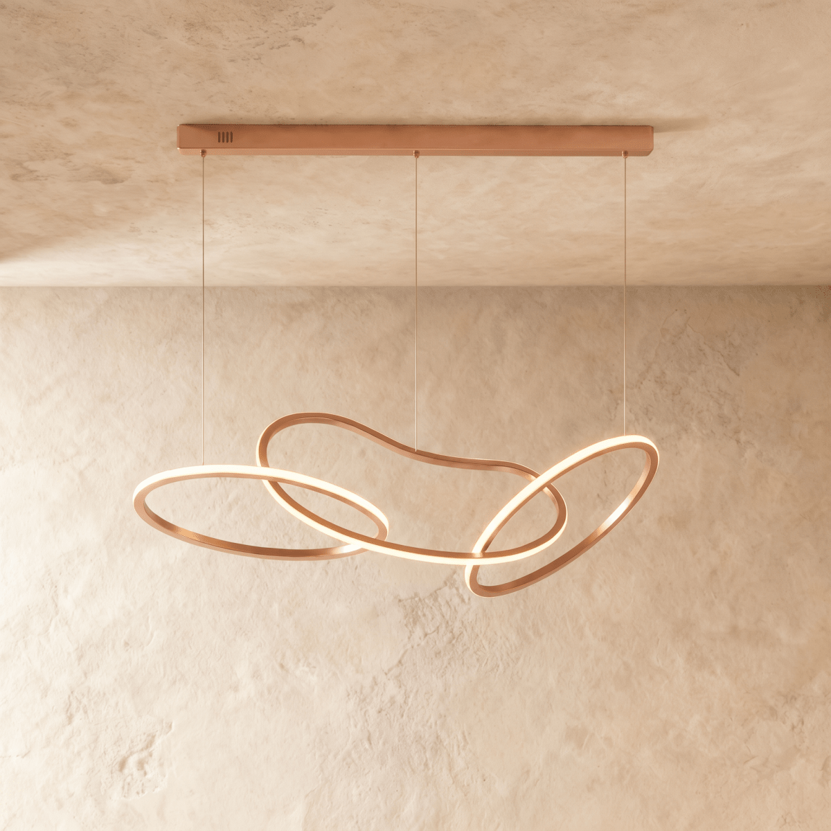 Clérachic chandelier in a warm rose gold finish, its looping rings glowing softly against a plaster wall.