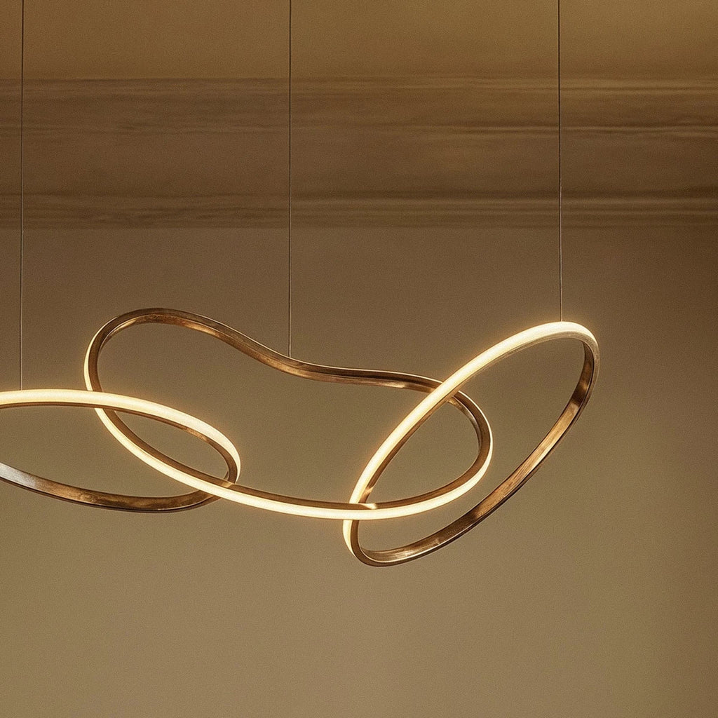 Detail of the Clérachic chandelier's interlocking gold loops, glowing warm white against a plaster wall and cornice.