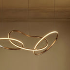 Detail of the Clérachic chandelier's interlocking gold loops, glowing warm white against a plaster wall and cornice.