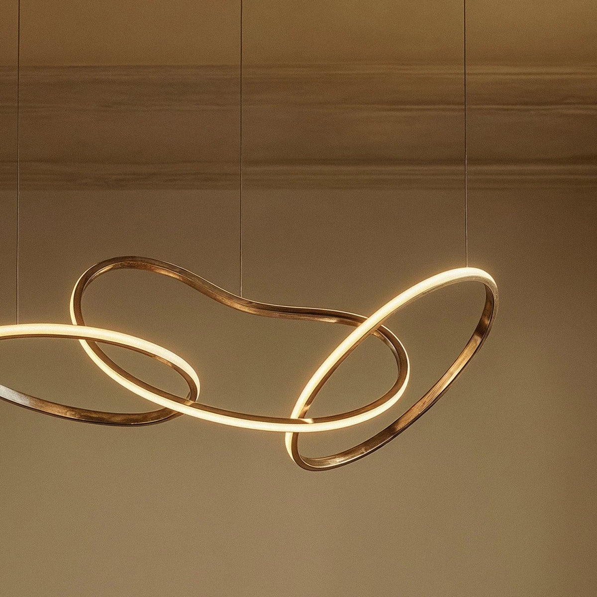 Detail of the Clérachic chandelier's interlocking gold loops, glowing warm white against a plaster wall and cornice.