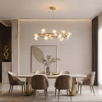 Round Clérachic chandelier with lit white ceramic petals above a dining table with brown chairs and a leaf-shaped wall panel.