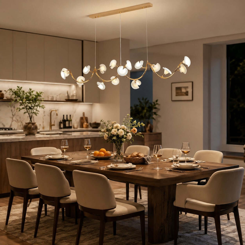 A Clérachic linear chandelier with petal shades hangs over a long set dining table, with an open kitchen lit behind it in the evening.