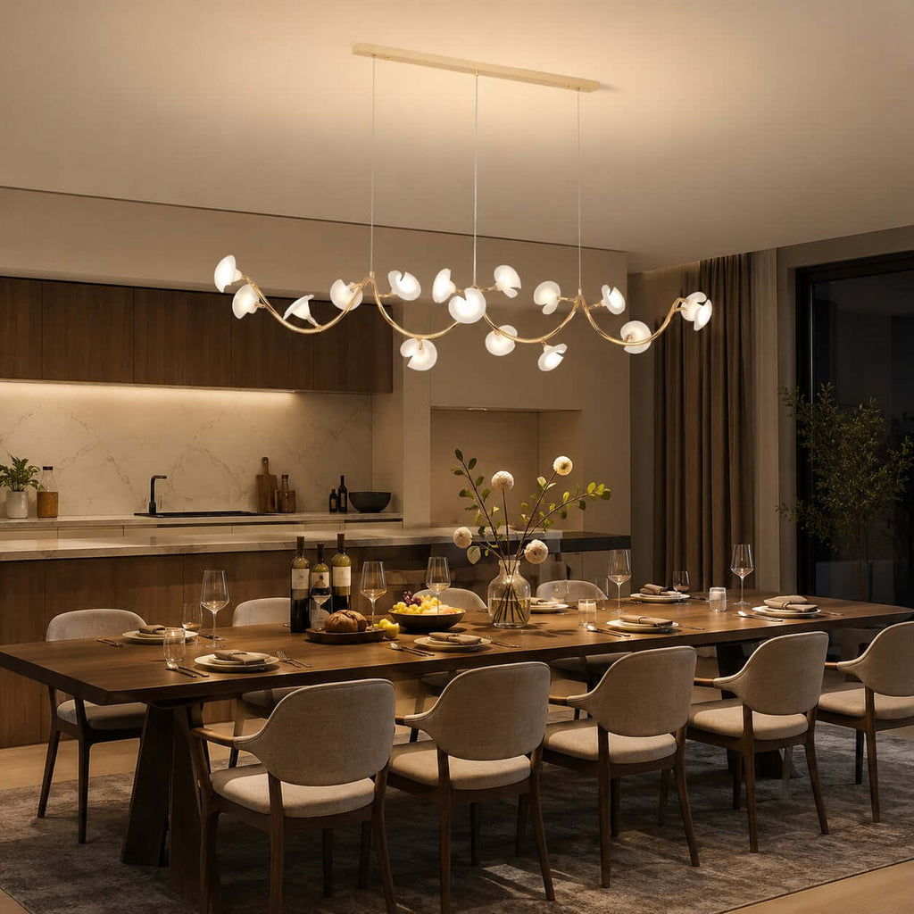 Evening view of a Clérachic linear chandelier running above a long wooden table laid for dinner, with a kitchen and curtains beyond.