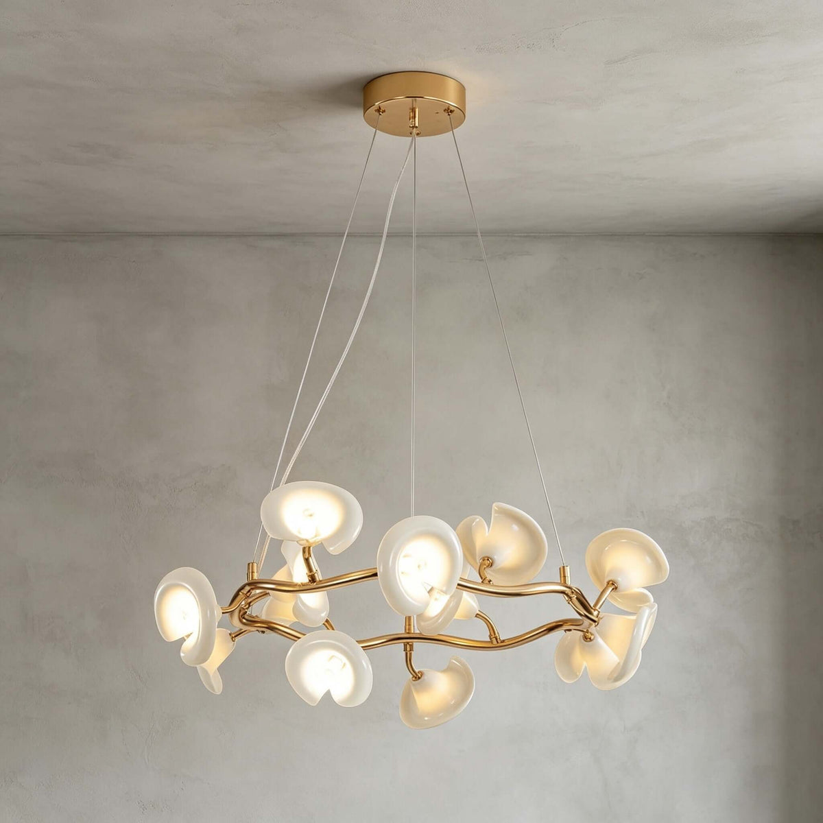 The round Clérachic chandelier hanging below a gold ceiling plate on thin wires, its ceramic petals lit against a grey limewash wall.