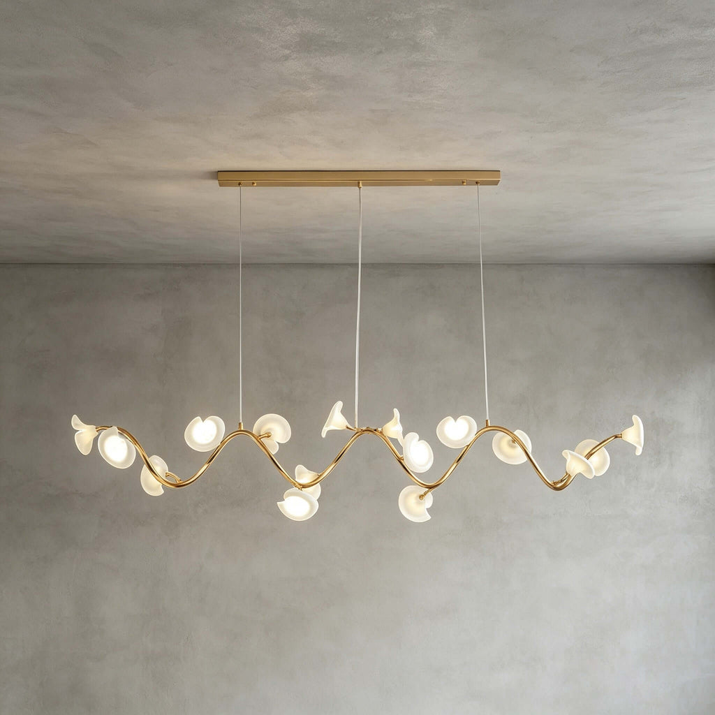 The linear Clérachic chandelier hangs from a long gold bar, its petals set along a wavy gold arm, against a grey wall.