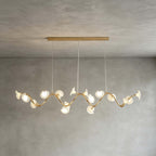 The linear Clérachic chandelier hangs from a long gold bar, its petals set along a wavy gold arm, against a grey wall.
