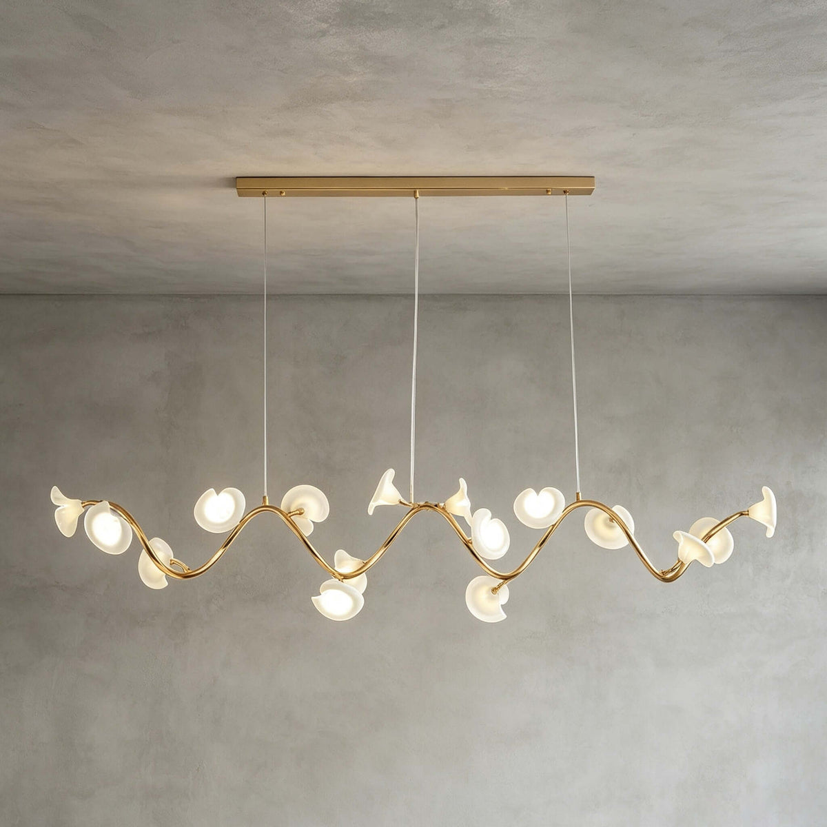 A Clérachic linear chandelier in its longer form, with petals along a wavy gold arm below a slim gold ceiling bar on grey limewash.