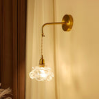 Lit warmly against cream curtains and a dark wood panel, this Clérachic rose-glass wall light hangs from a curved brass arm.