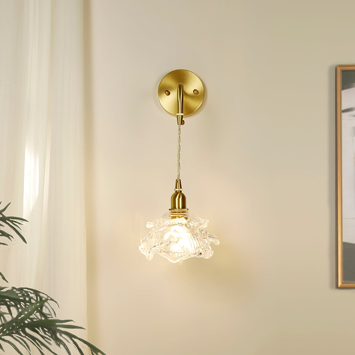 By a cream curtain and a framed picture, this Clérachic wall light hangs its small glass rose shade from a round brass plate.
