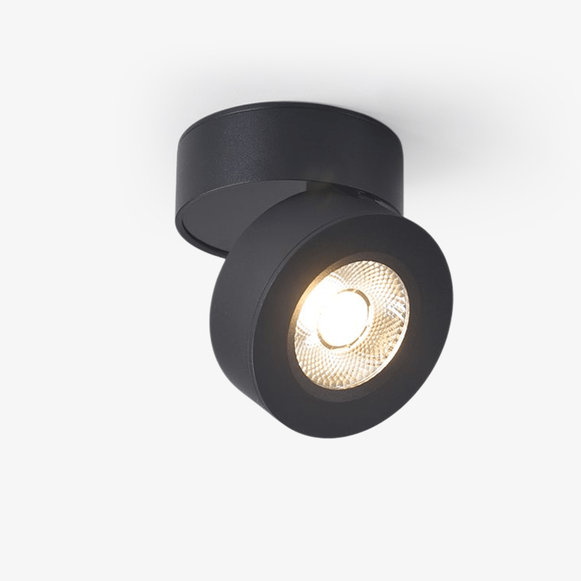This Clérachic round spotlight in black on a plain light backdrop, with its tilted disc facing out and a warm LED glowing in a ribbed reflector.