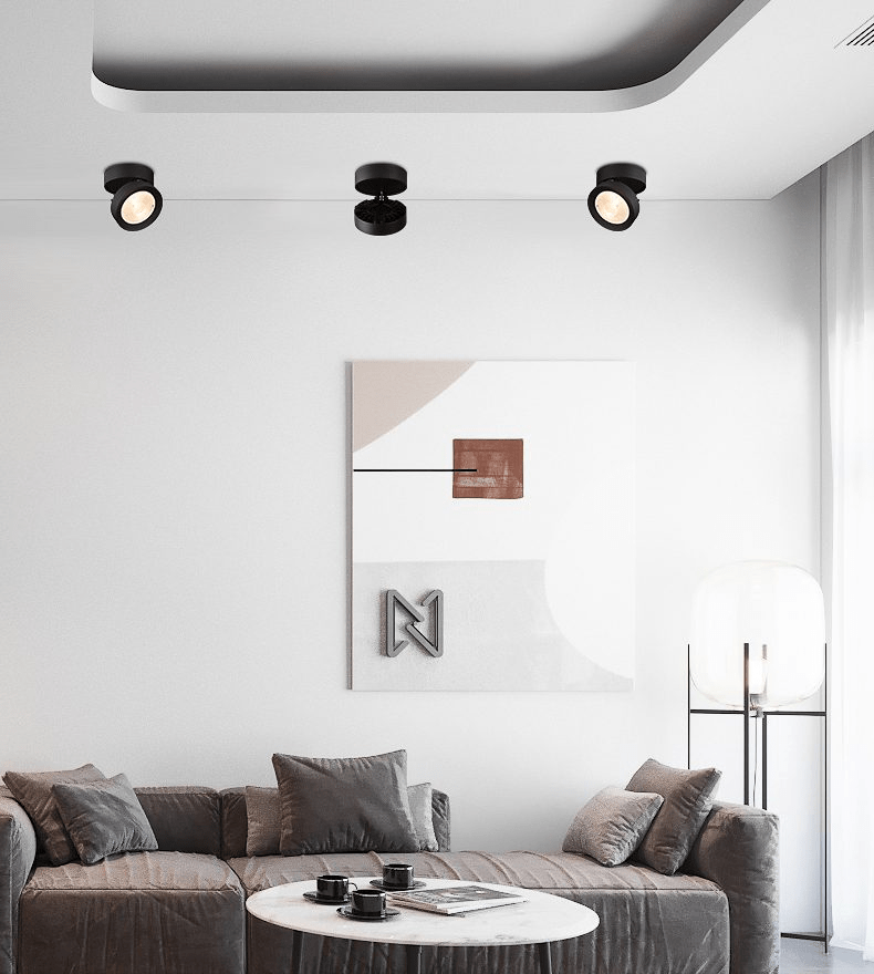Three black Clérachic spotlights along a ceiling line in a living room, above a grey sofa, a round white coffee table and an abstract print.