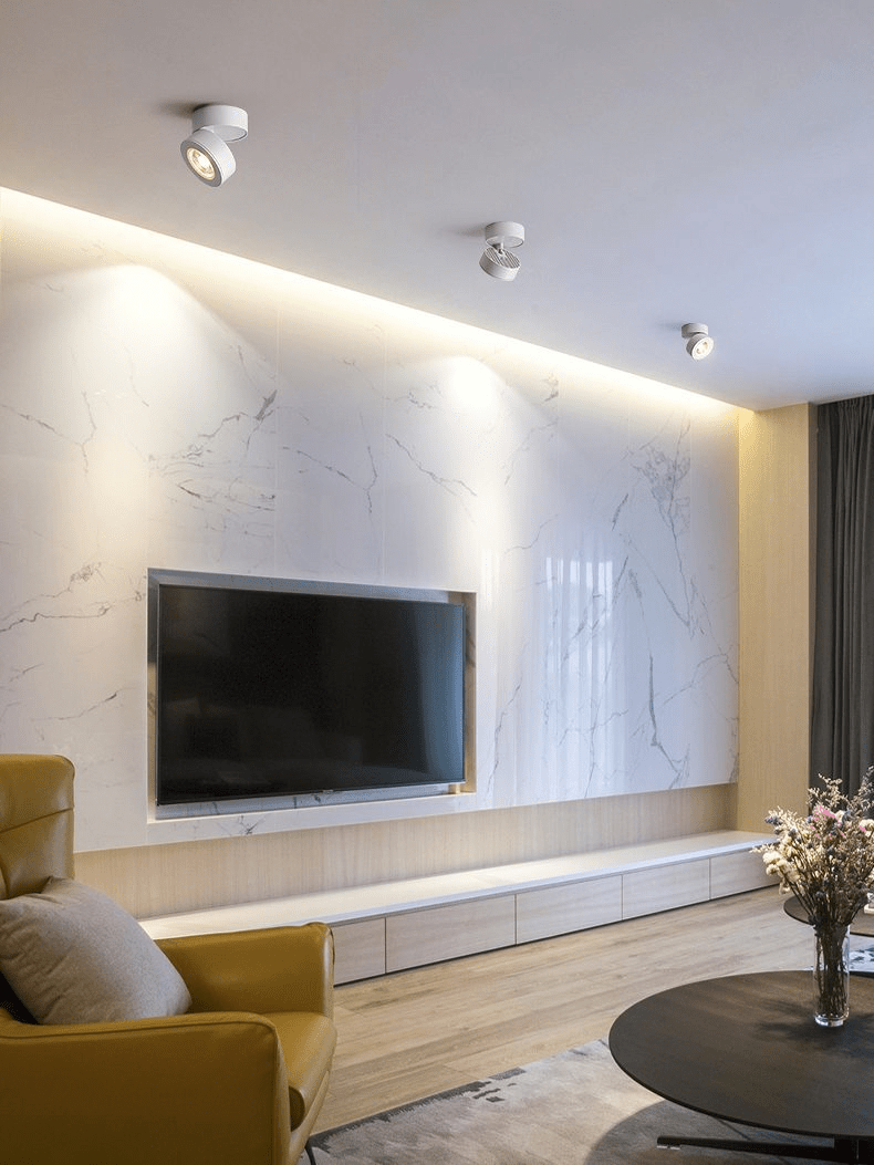 White Clérachic spotlights on the ceiling of a living room with a marble-look feature wall, a wall-mounted television and a mustard armchair.