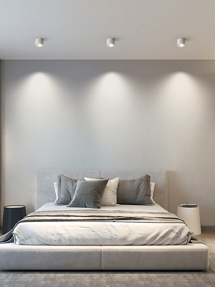 Three white Clérachic spotlights on a bedroom ceiling, each throwing a soft cone of light down the wall above a low grey bed.
